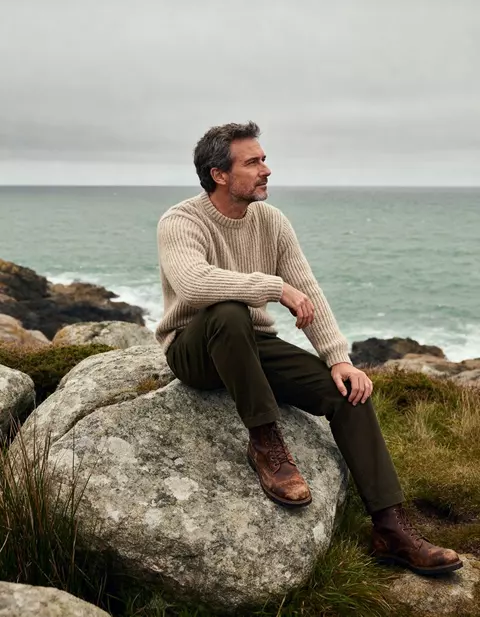 Hombre con jersey de punto sentado en una roca junto al mar